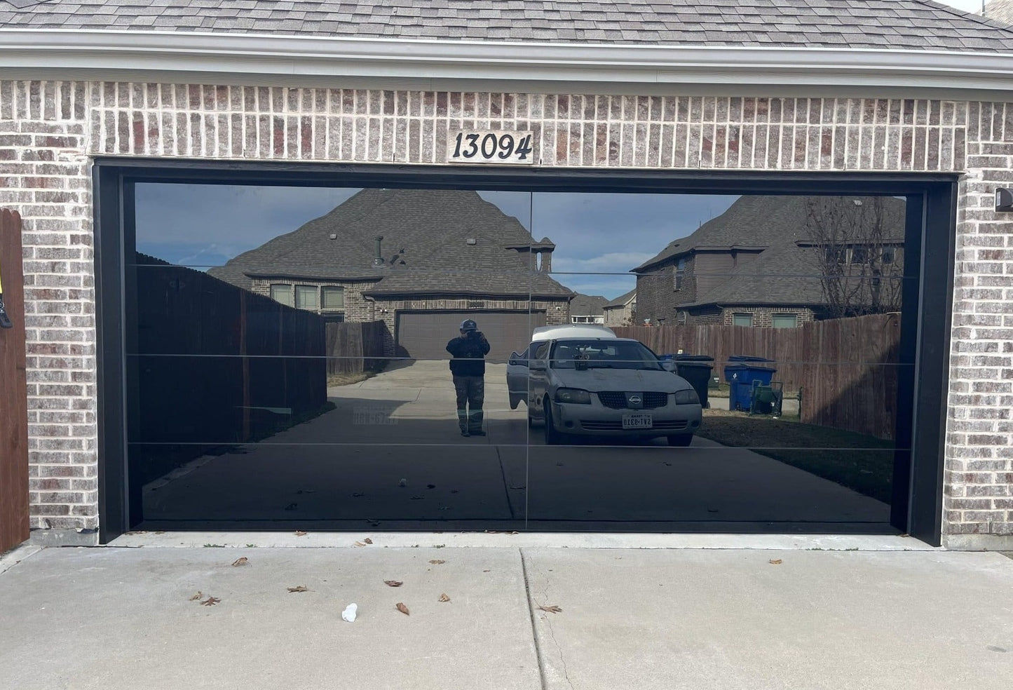 Modern black-tinted glass garage door installed on a brick house, reflecting the driveway and surrounding neighborhood, showcasing the sleek and contemporary design