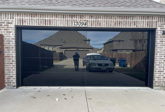 Modern black-tinted glass garage door installed on a brick house, reflecting the driveway and surrounding neighborhood, showcasing the sleek and contemporary design