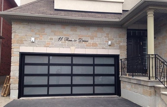Elegant residential garage door featuring a matte black frame with frosted glass panels, set against a textured stone facade of a home, enhancing the property's curb appeal and contemporary design