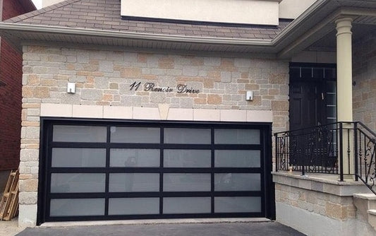 Elegant residential garage door featuring a matte black frame with frosted glass panels, set against a textured stone facade of a home, enhancing the property's curb appeal and contemporary design
