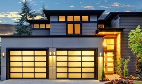 Stunning modern residence at dusk with two illuminated garage doors featuring frosted glass panels in a matte black frame, beautifully complementing the dark grey and wooden exterior of the luxury home
