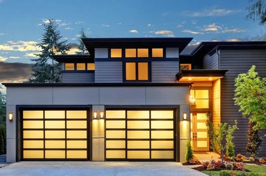 Stunning modern residence at dusk with two illuminated garage doors featuring frosted glass panels in a matte black frame, beautifully complementing the dark grey and wooden exterior of the luxury home